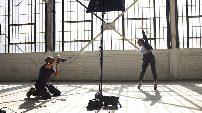 Photographer kneeling while shooting a model posing under studio lighting in an industrial space.
