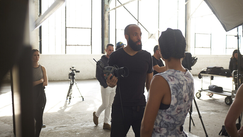 Director demonstrating camera technique to crew members on a bright, industrial film set.