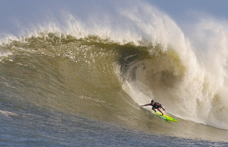 Surfer in wetsuit riding a massive wave with white foam crest and churning water.
