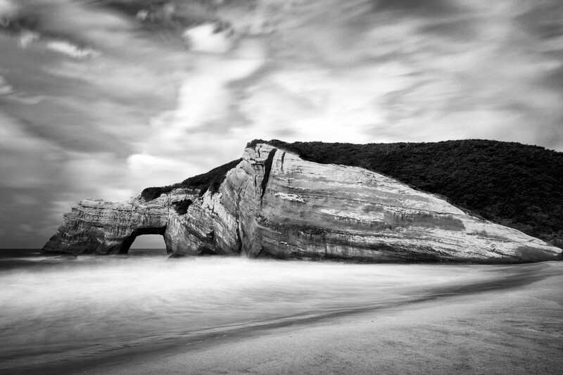 Weathered rock formation on a beach with a natural arch, captured in black and white photography.