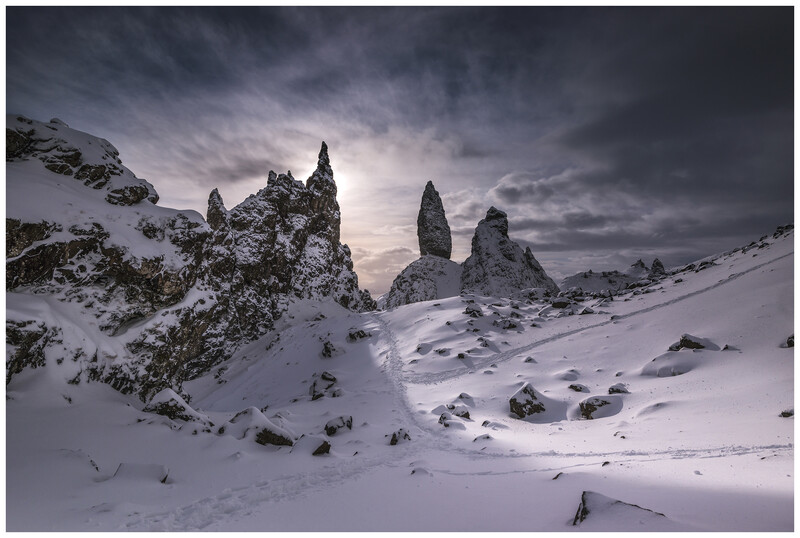 Snow-covered landscape with distinctive rock formations rising from a frozen valley under stormy clouds.