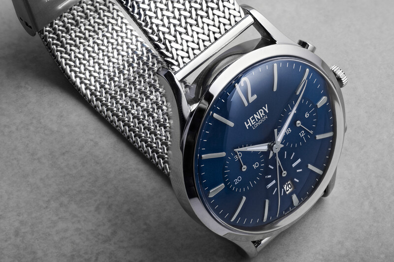 Luxury chronograph watch with navy dial and mesh metal band displayed against gray background.