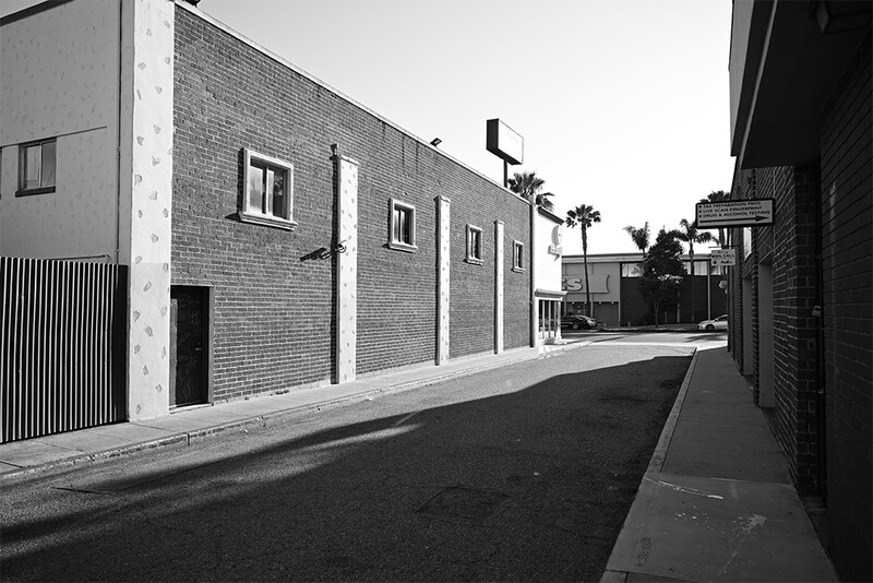 Brick building facade along empty urban alley with palm trees visible in distance.