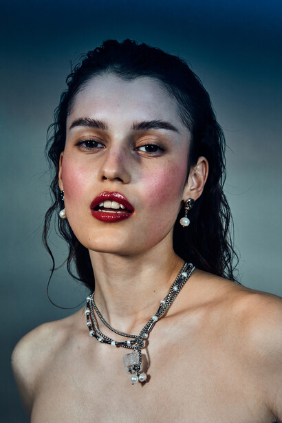 Portrait of a woman with dark wet-look hair, red lipstick, and a statement crystal necklace against a teal background.