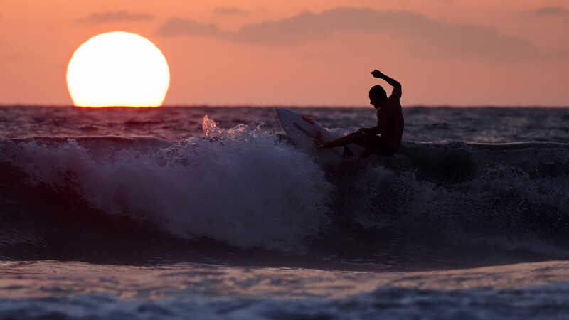 Silhouetted surfer riding a wave during golden hour sunset with large sun on horizon.