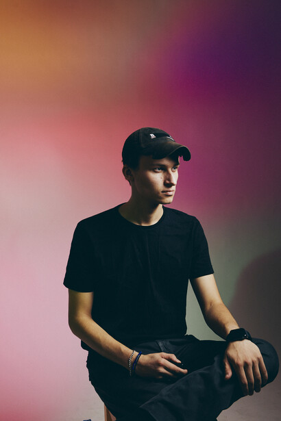 Person wearing black cap and t-shirt against gradient pink and purple background.
