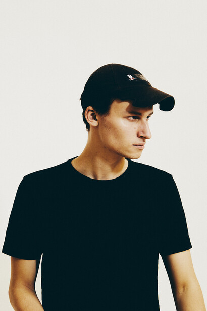 Man wearing black t-shirt and dark baseball cap, looking to the side against neutral background.