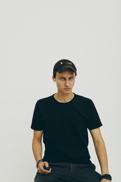 Young man in dark t-shirt and cap against neutral background, looking directly at camera.