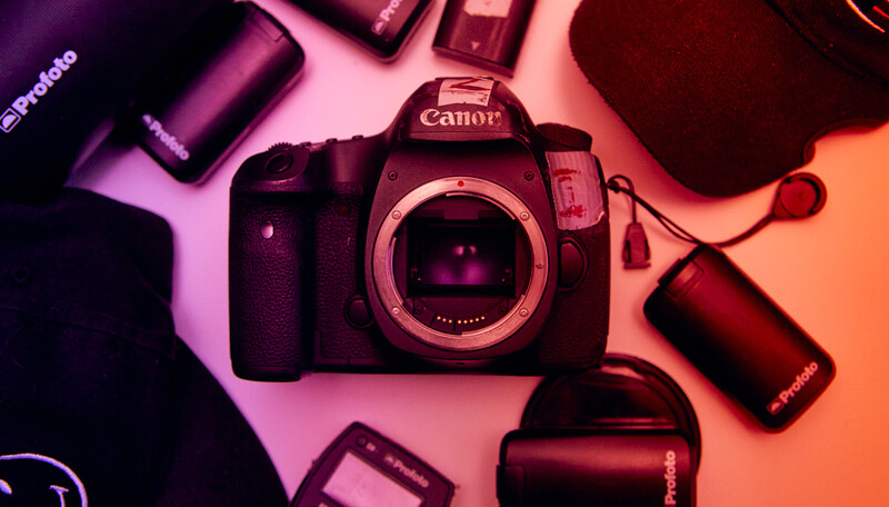 Canon DSLR camera body surrounded by batteries and accessories on a pink and orange gradient background.