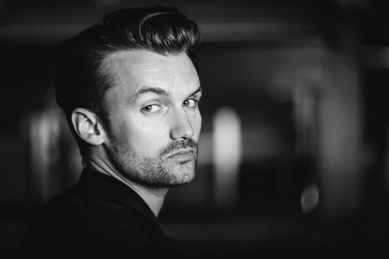 Man with styled dark hair and facial hair looking over shoulder in black and white portrait.