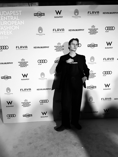 Person in black clothing standing against a branded step-and-repeat backdrop at a fashion event.