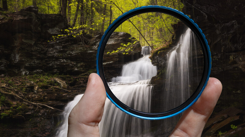 Hand holding a circular filter frame showing a tiered waterfall in soft focus through the filter.