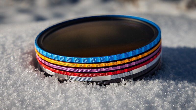 Stacked circular lens filters with colored fabric bands in blue, yellow, red, and black on snow.