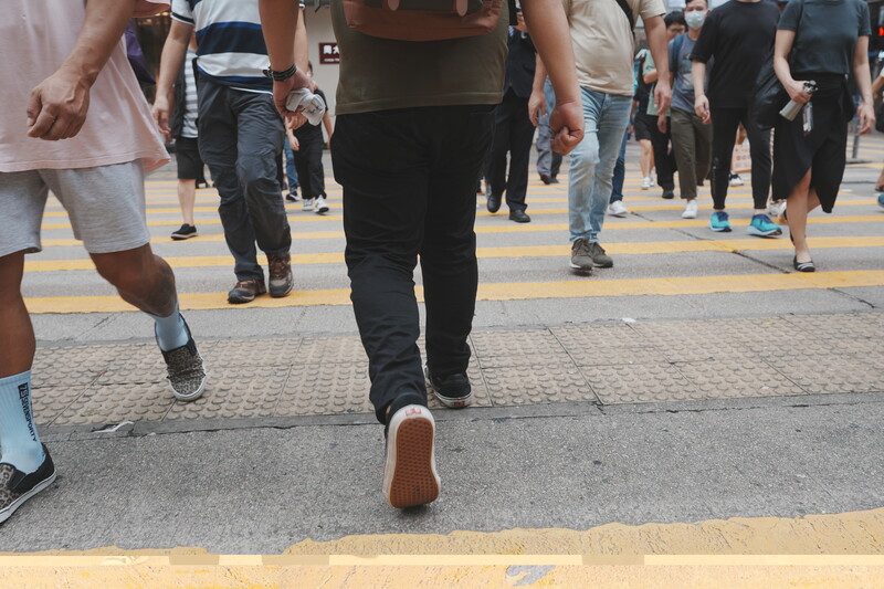 Runner's shoe captured mid-stride on a paved crosswalk surrounded by pedestrians.