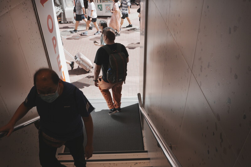 People walking through a narrow white corridor with bright daylight visible at the exit.