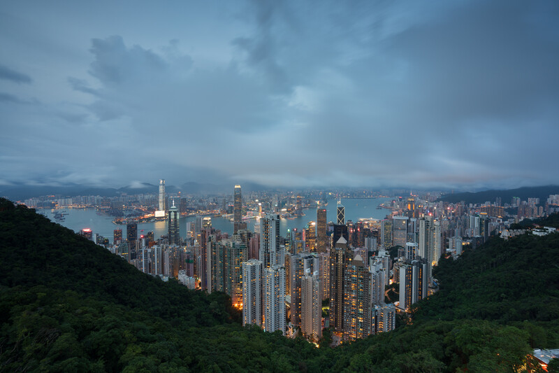 Dense urban skyline at dusk with illuminated skyscrapers framed by forested hillsides.
