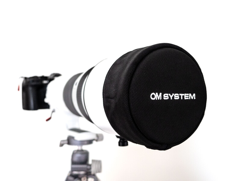 OM System telephoto lens with lens cap and tripod collar mounted on ball head tripod.