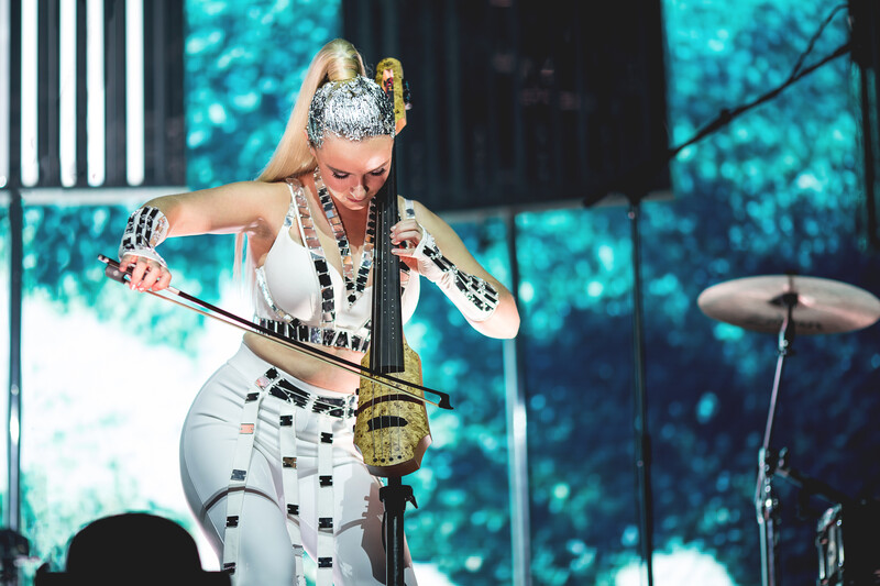 Performer in white sequined outfit and sparkling crown plays saxophone on stage with turquoise lighting backdrop.