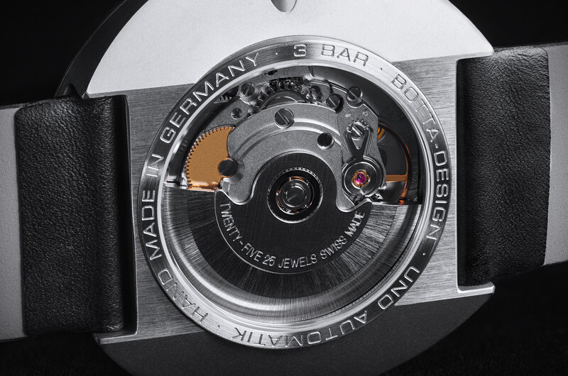 Close-up of a watch case back showing mechanical movement with orange accents and German manufacturing text.