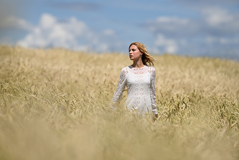 Woman in white lace dress standing in a golden grain field under a partly cloudy sky.