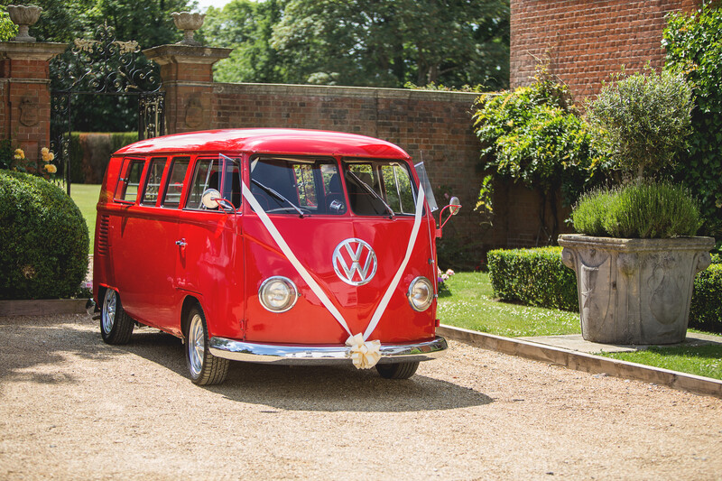 Bright red Volkswagen Type 2 bus parked on gravel in a manicured garden setting.