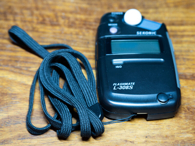 Sekonic Flashmate L308S light meter with blue fabric strap coiled beside it on wooden surface.