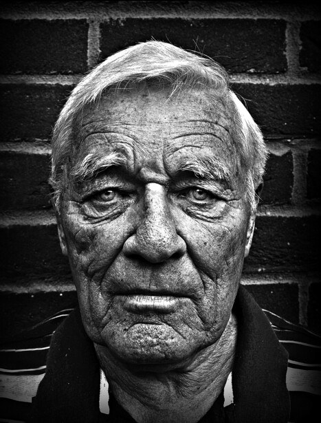 Black and white portrait of an older man with glasses against a brick wall.