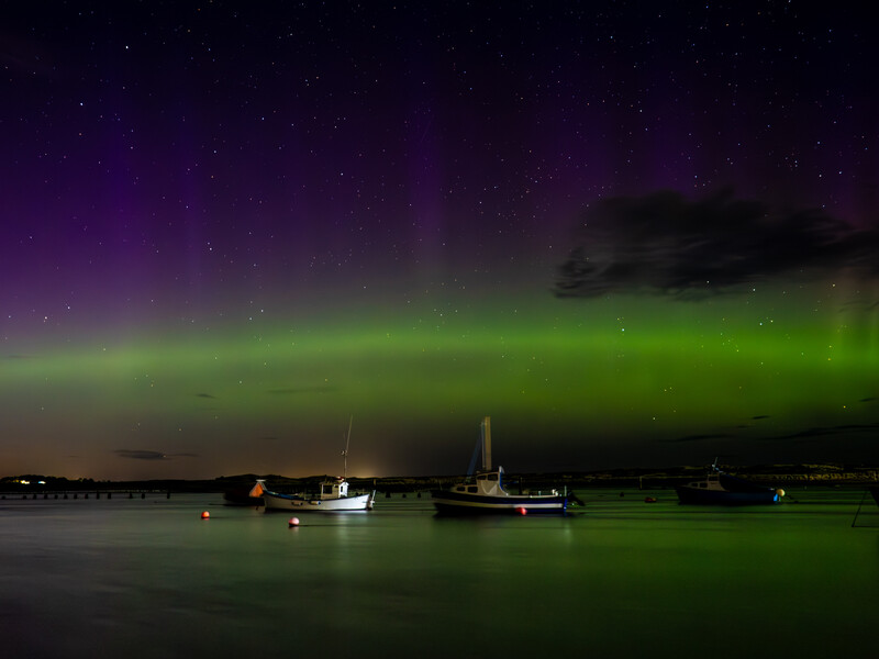 Aurora borealis illuminates night sky above moored sailboats on calm water.