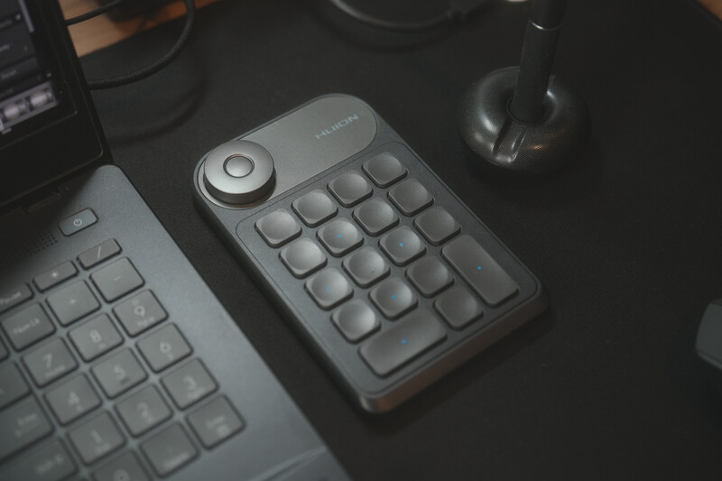 Gray mechanical keypad with rotary encoder knob positioned on dark desk surface next to keyboard.