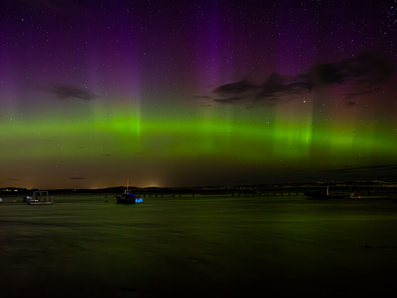 Aurora borealis illuminates night sky with green and purple bands above dark water and silhouetted boats.