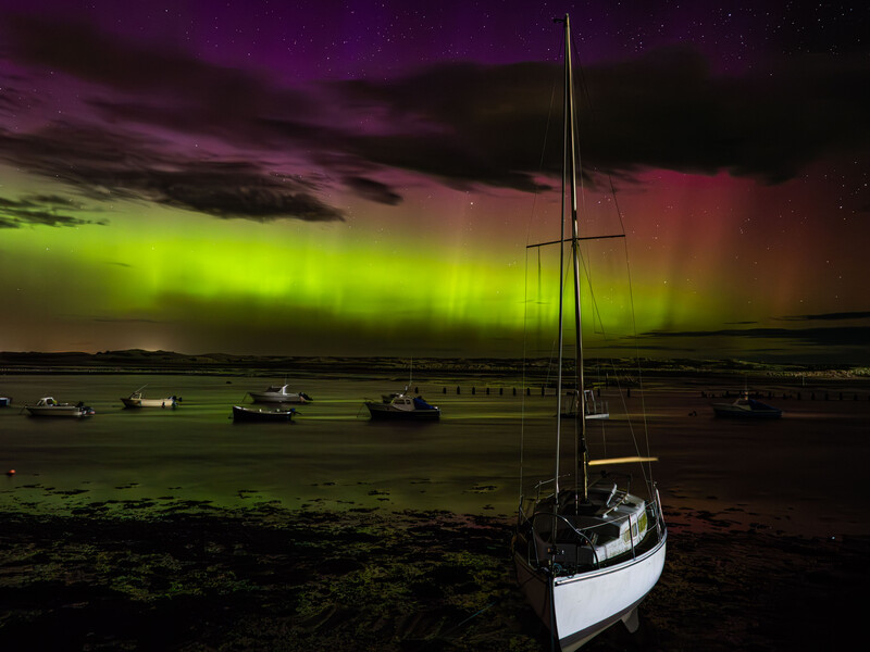 Sailboat anchored in shallow water beneath a vibrant aurora borealis display in purple and green hues.