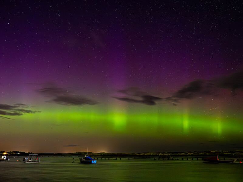 Aurora borealis glowing green and bright yellow across the night sky above a dark landscape with buildings and boats.