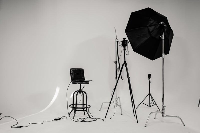 Professional photography studio setup with lighting equipment including an umbrella reflector, light stand, and monitor on an industrial stool against a white backdrop.