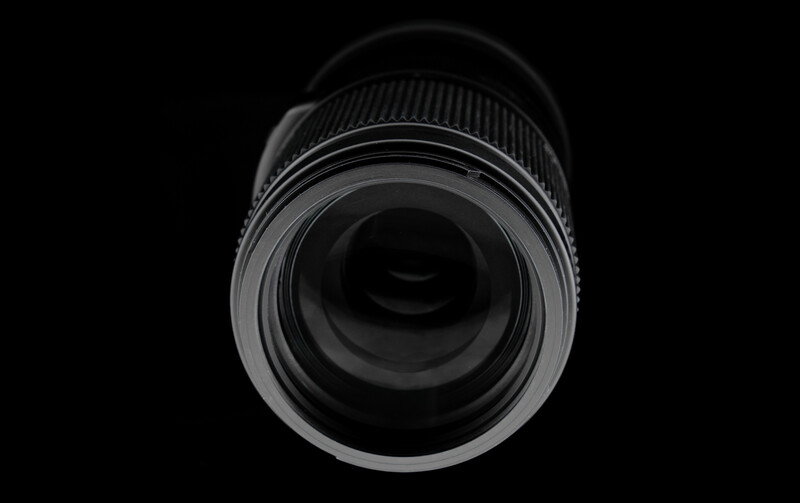 Close-up front view of a camera lens against a black background, showing internal glass elements and focusing ring.