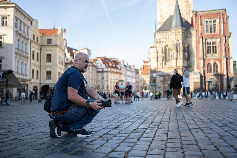 Photographer crouching in a European town square with a camera, surrounded by historic architecture and pedestrians.