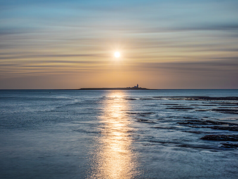 Sun reflecting on calm ocean water during golden hour sunset with distant silhouetted landmass on horizon.