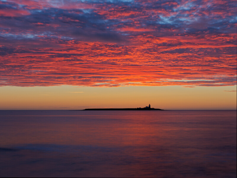 Silhouetted lighthouse on the horizon beneath a dramatic crimson and orange sunset sky reflected in calm water.