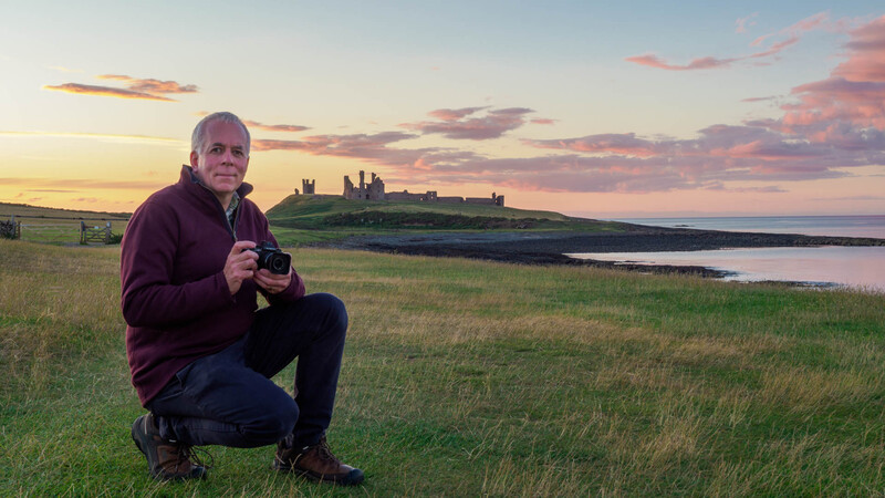 Photographer crouching in a grassy field at golden hour, holding a camera, with a coastal castle ruins visible on a distant hilltop.