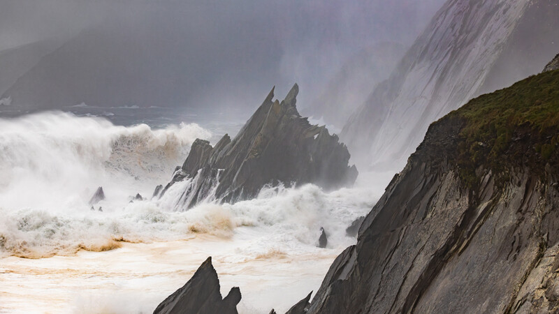 Dramatic seascape with jagged rock formations surrounded by powerful white waves and sea spray.