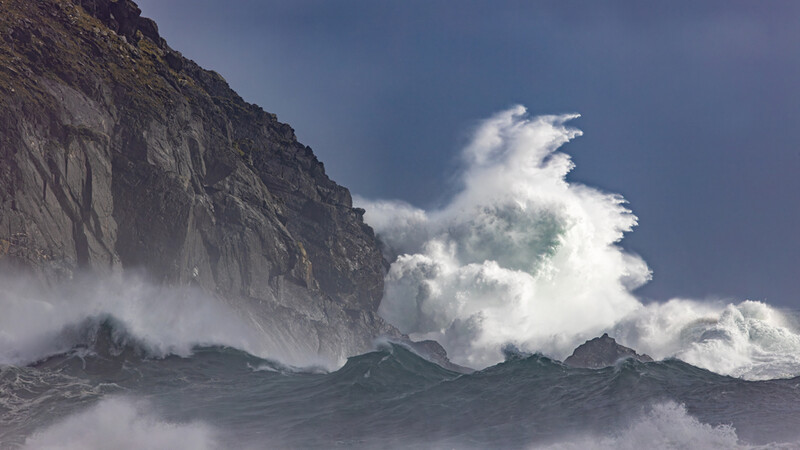 Massive ocean waves crash against a steep rocky cliff under a dramatic blue sky with white clouds.