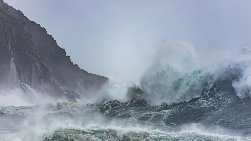 Massive waves with seafoam crash against a rocky cliff during a dramatic storm at the coastline.