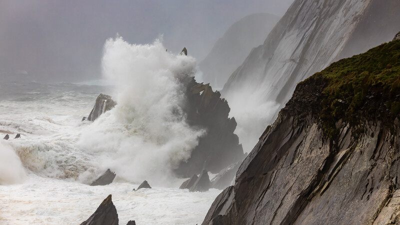 Massive ocean waves crash against rocky cliffs shrouded in sea spray and mist.