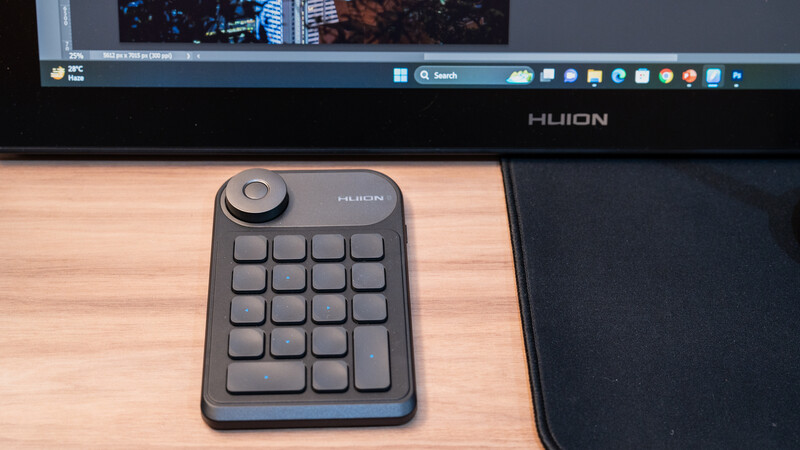 Huion keypad with 16 programmable buttons and rotary dial on wooden desk next to monitor.