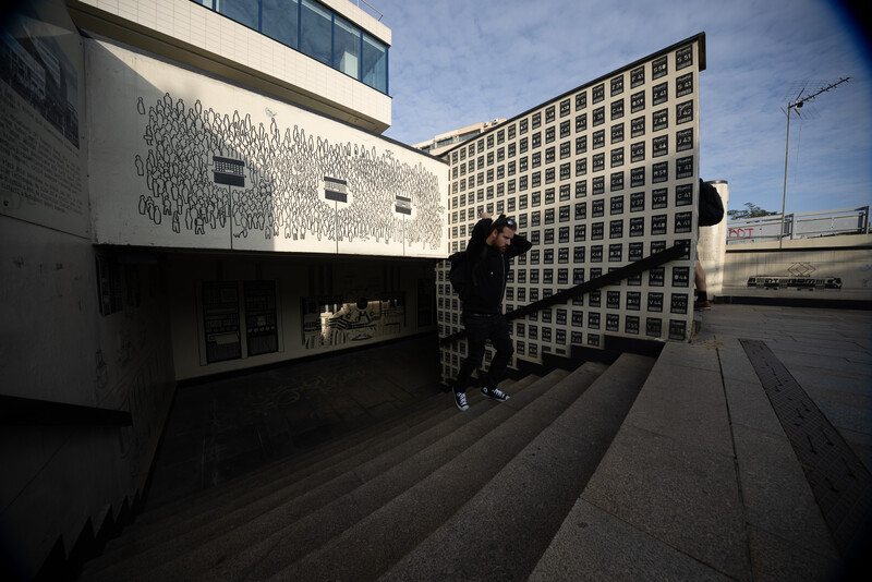 Two people standing in shadow between modern white and grid-patterned buildings on a sunlit plaza.