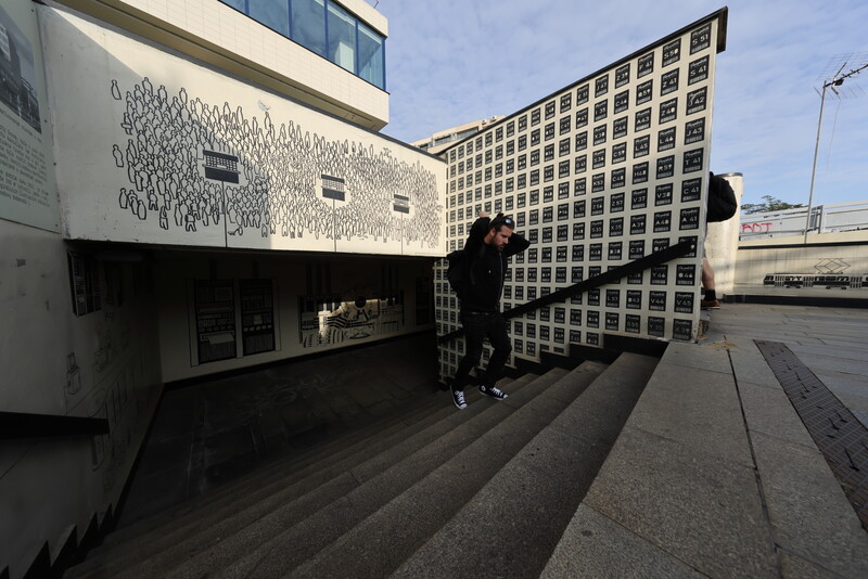 Person walking through modern urban courtyard with geometric architecture and angular building facades.
