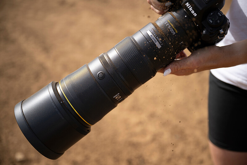 Telephoto lens with gold accents mounted on a camera body, held at an angle against an outdoor background.