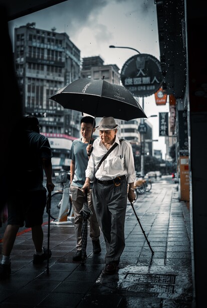 Two men in formal attire standing under a large black umbrella on a wet city street.