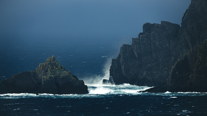 Dramatic coastal rock formations with powerful waves crashing against dark cliffs under stormy skies.