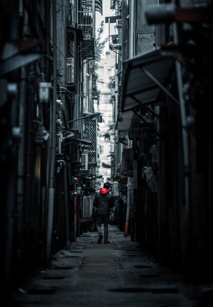 Person in red jacket walking alone through narrow dark urban alley with tall buildings.