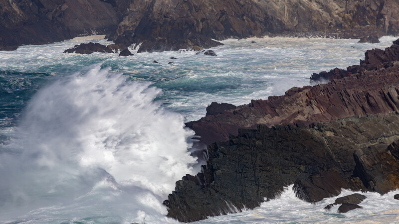 Dramatic coastal waves crash against dark columnar basalt rock formations jutting from turbulent turquoise water.